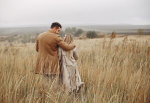 Couple walking arm in arm through a tall grassy field, viewed from behind on an overcast day.