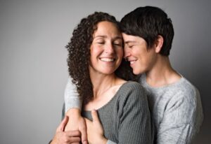 Two women smiling with their eyes closed while embracing against a neutral grey background.