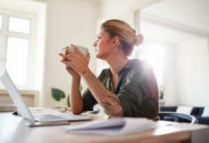Person sitting at a table with a laptop, holding a mug and looking thoughtfully toward a bright window.