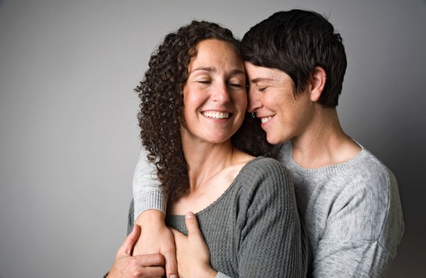 Two women embracing closely and smiling against a plain grey background.